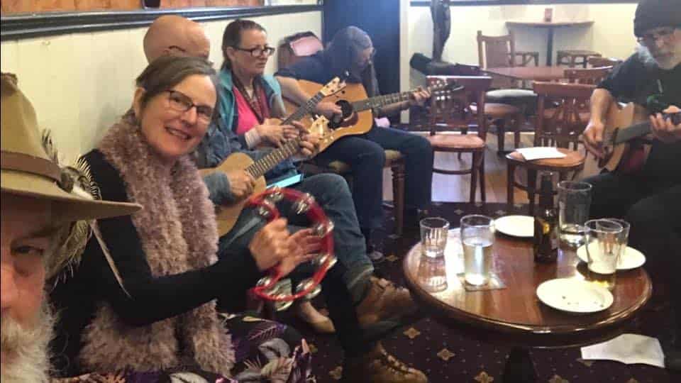 Raw Jam wiht musicians playing a guitars and a tambourine while sitting on a side bench at the pub.